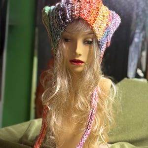 Könnte beinhalten: Ein handgefertigter, farbenfroher Häkelhut mit einem Regenbogenverlauf aus Farben, darunter Orange, Gelb, Grün, Blau und Lila. Der Hut hat eine einzigartige Form und wird auf einem Schaufensterkopf präsentiert. Ein passender Häkelbinder hängt herunter.