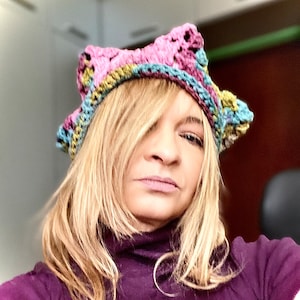 May include: A woman wearing a purple turtleneck shirt and a multi-colored crocheted crown.