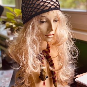 Puede incluir: Una cabeza de maniquí con cabello rubio, que lleva una gorra de red negra y una bufanda de serpiente de ganchillo. La serpiente es verde oliva y burdeos con ojos grandes y una lengua roja. El fondo está borroso.