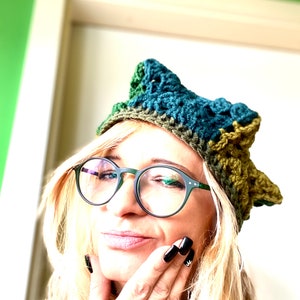 May include: A woman wearing a green, blue, and brown crocheted hat with a pointed top. She is also wearing black framed glasses and has black nail polish on her nails.