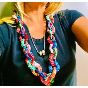 May include: A chunky necklace featuring large, interlocking circular links crocheted in a vibrant multicolored yarn pattern of red, blue, green, and yellow. A secondary thin silver-toned metal chain with two small cylindrical beads hangs underneath.