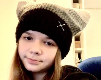 Könnte beinhalten: Eine Person trägt eine schwarze und graue Strickmütze mit einem weißen X, das auf die Vorderseite gestickt ist.