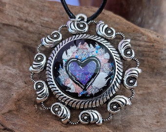 Könnte beinhalten: Eine silberne Halskette mit einem runden Anhänger aus schwarzem und silbernem Rahmen. Der Rahmen hat einen runden, transparenten Glaseinsatz mit einem violetten Herzen darin. Das Herz hat eine glitzernde, irisierende Oberfläche.