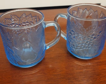 May include: Two blue glass mugs with a grape and leaf design. The mugs have a handle and are made of clear glass with a frosted blue finish.