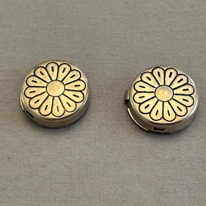 May include: Two small, round, silver-colored containers with a floral design on the top. The flower has a central circle with petal-like shapes around it. The containers are on a gray surface.