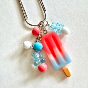 May include: A silver metal paperclip charm featuring a miniature popsicle pendant with a red, white, and blue gradient. The charm is decorated with small round beads in red, white, and blue, along with two translucent light blue bear-shaped beads.