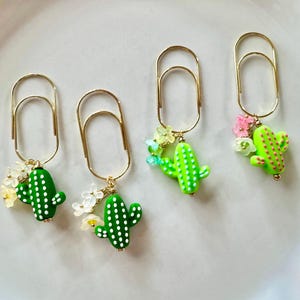 May include: Four gold-toned metal paper clips, each featuring a dangling charm of a green cactus with white or pink dots. Each cactus charm is accompanied by small, translucent, multi-colored flower-shaped beads attached with gold-toned jump rings.