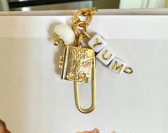 May include: A gold-colored charm featuring a miniature cookbook, a white bead, and letter beads spelling "YUM". The cookbook charm has the words "COOK BOOK" on the cover. The charm is attached to a gold-colored paperclip.