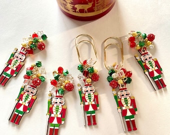 May include: A collection of Christmas-themed paper clips featuring a nutcracker design. Each clip is adorned with red, green, and gold beads. A red and gold decorative container is in the background.