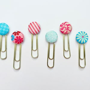 May include: Six gold paper clips with colorful fabric-covered buttons. The buttons feature various patterns, including stripes, floral designs, and geometric shapes. The buttons are in shades of blue, pink, and white.