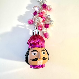 Op de afbeelding: Een roze kerstversiering in de vorm van een notenkrakersoldaat, versierd met een kroon en een snor. Het is versierd met een cluster van roze en zilveren kralen en bevestigd aan een zilveren sluiting. Feestelijke decoratie.