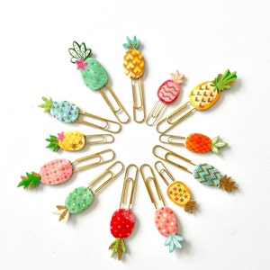 May include: A set of 13 gold paper clips with pineapple designs. Each clip features a different pineapple pattern with various colours, including green, yellow, pink, blue, and orange.