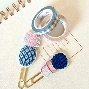 Puede incluir: Dos clips de papel dorados con diseños bordados de piña y cactus. La piña es azul y rosa, y el cactus es azul y rosa con una maceta rosa. Los clips están en una libreta rosa con una espiral dorada.