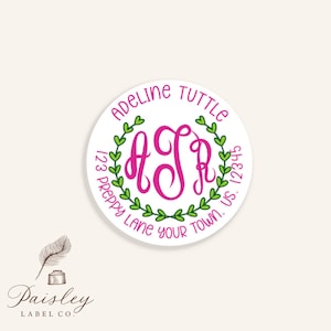 May include: A round white sticker with a pink monogram inside a green leaf wreath. The text "Adeline Tuttle" and address details are in a circular pattern around the monogram. The sticker is from Paisley Label Co.