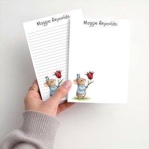 May include: Two white stationery notepads featuring the name Maggie Reynolds printed at the top. One notepad is lined, and the other is blank. Both feature a small illustration of a brown mouse in a blue sweater holding a red tulip.