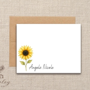 May include: White stationery card with a watercolor sunflower illustration and the name "Angela Nicole" in script. The card is paired with a brown envelope, set against a neutral linen background.