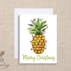 May include: A watercolor Christmas card featuring a pineapple decorated with colorful Christmas lights. The card has the words "Merry Christmas" in green script. The card is on a white background.