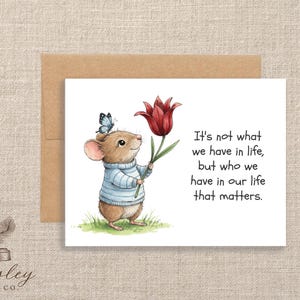 May include: Greeting card featuring a charming illustration of a mouse wearing a blue and white striped sweater, holding a red tulip and a butterfly. The card includes the text: "It's not what we have in life, but who we have in our life that matters."