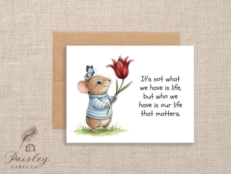 May include: Greeting card featuring a charming illustration of a mouse wearing a blue and white striped sweater, holding a red tulip and a butterfly. The card includes the text: "It's not what we have in life, but who we have in our life that matters."