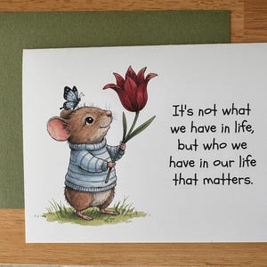 May include: Greeting card featuring a charming illustration of a mouse wearing a blue sweater and holding a red tulip. A blue butterfly rests on the mouse's head. The card includes the text: "It's not what we have in life, but who we have in our life that matters."