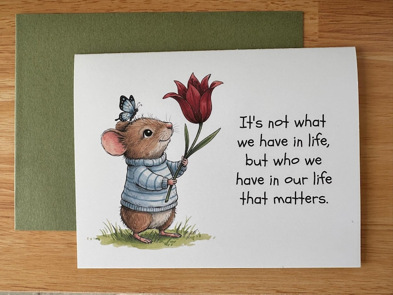 May include: Greeting card featuring a charming illustration of a mouse wearing a blue sweater and holding a red tulip. A blue butterfly rests on the mouse's head. The card includes the text: "It's not what we have in life, but who we have in our life that matters."