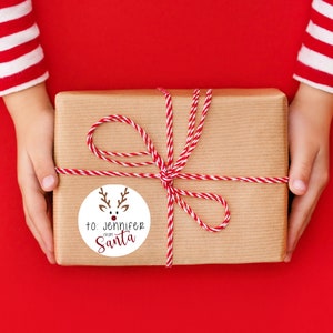 Puede incluir: Una caja de regalo marr&oacute;n envuelta con cordel rojo y blanco y una etiqueta de regalo redonda con una ilustraci&oacute;n de reno. La etiqueta dice "para: Jennifer de Santa Claus".