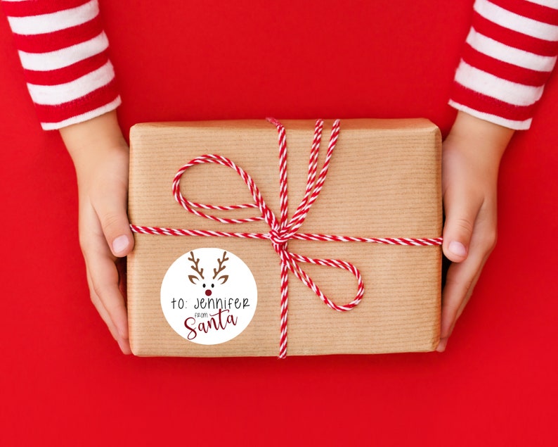 Puede incluir: Una caja de regalo marr&oacute;n envuelta con cordel rojo y blanco y una etiqueta de regalo redonda con una ilustraci&oacute;n de reno. La etiqueta dice "para: Jennifer de Santa Claus".