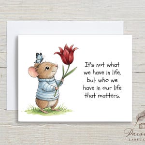 May include: Greeting card featuring a watercolor illustration of a mouse wearing a blue sweater, holding a red tulip, and a butterfly on its head. The card includes the text: "It's not what we have in life, but who we have in our life that matters."
