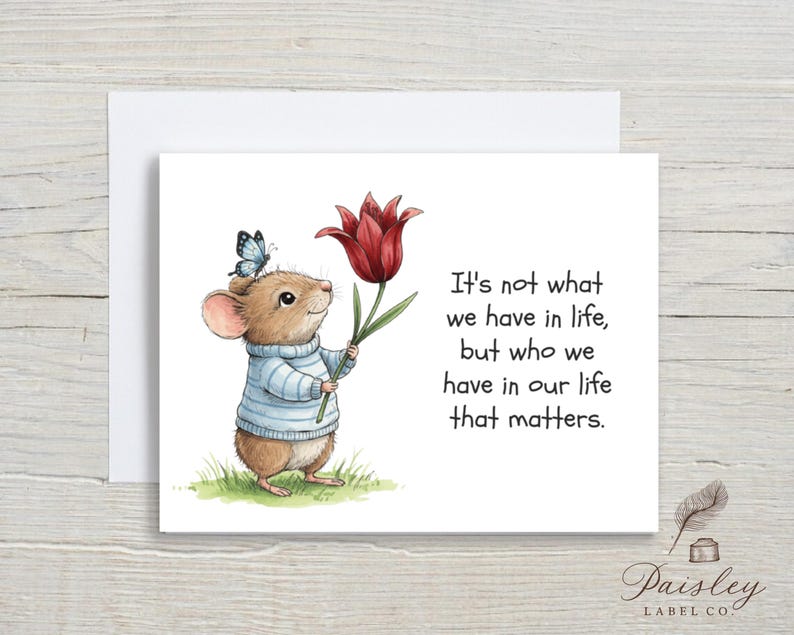 May include: Greeting card featuring a watercolor illustration of a mouse wearing a blue sweater, holding a red tulip, and a butterfly on its head. The card includes the text: "It's not what we have in life, but who we have in our life that matters."