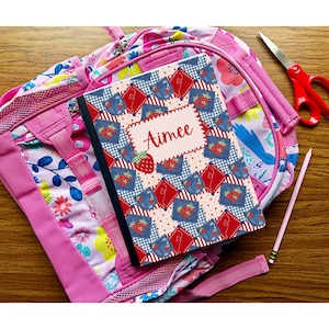May include: A pink backpack with floral patterns and a notebook. The notebook has a strawberry and patchwork design with the name "Aimee" on it. A pair of red scissors and a pencil are also visible.