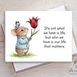 May include: Greeting card featuring a watercolor illustration of a mouse wearing a blue sweater and holding a red tulip. A blue butterfly rests on the mouse's head. The card includes the text: "It's not what we have in life, but who we have in our life that matters."
