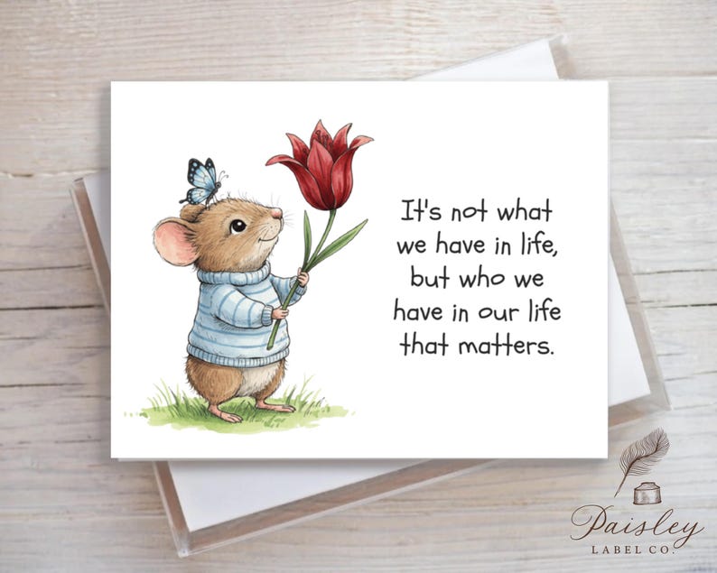 May include: Greeting card featuring a watercolor illustration of a mouse wearing a blue sweater and holding a red tulip. A blue butterfly rests on the mouse's head. The card includes the text: "It's not what we have in life, but who we have in our life that matters."