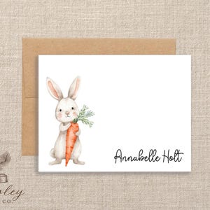 May include: A white card with a watercolor illustration of a bunny holding a carrot. The bunny has pink ears and is holding a large orange carrot with green leaves. The name "Annabella Holt" is written in black script.