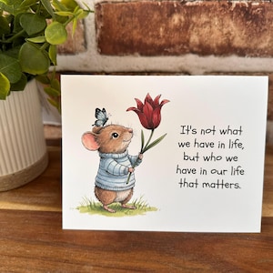 May include: A greeting card featuring a cartoon mouse wearing a blue sweater and holding a red tulip. A butterfly rests on the mouse's head. The card has the text: "It's not what we have in life, but who we have in our life that matters."