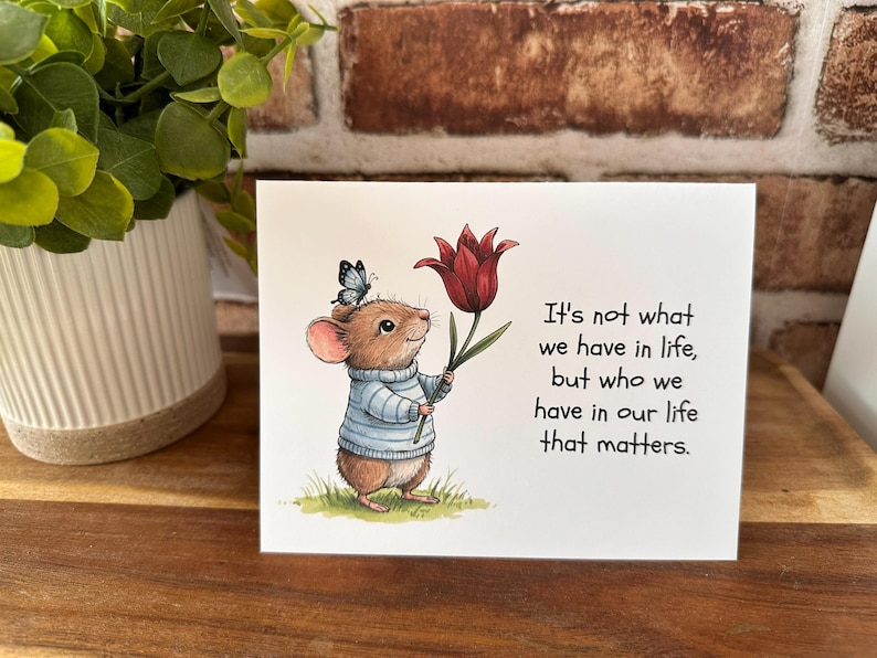 May include: A greeting card featuring a cartoon mouse wearing a blue sweater and holding a red tulip. A butterfly rests on the mouse's head. The card has the text: "It's not what we have in life, but who we have in our life that matters."
