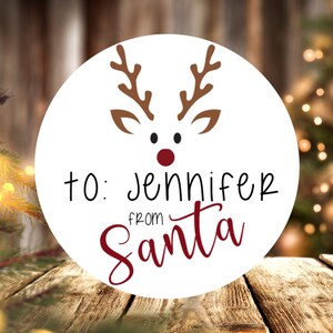 Puede incluir: Etiqueta de regalo redonda blanca con una ilustraci&oacute;n de un reno con la nariz roja. La etiqueta dice "Para: Jennifer de Santa Claus" en letras negras y rojas.