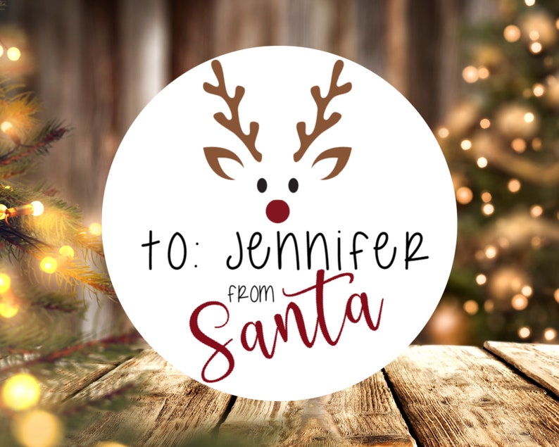 Puede incluir: Etiqueta de regalo redonda blanca con una ilustraci&oacute;n de un reno con la nariz roja. La etiqueta dice "Para: Jennifer de Santa Claus" en letras negras y rojas.