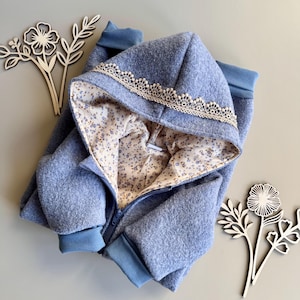 Könnte beinhalten: Hellblaue Kapuzenjacke mit floral gemustertem Futter und cremefarbenem Spitzenbesatz an der Kapuze. Die Jacke hat einen durchgehenden Reißverschluss und blaue Rippbündchen. Dekorative Blumen aus Holz an den Seiten.