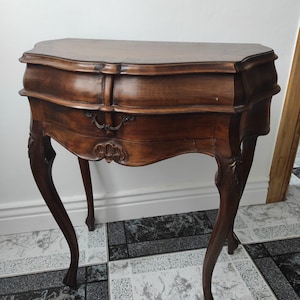Op de afbeelding: Een donkerbruine antieke houten bijzettafel met een gebogen voorkant en sierlijke details. De tafel heeft een enkele lade met een decoratieve handgreep en vier gebogen poten. De bovenkant van de tafel is lichtbruin.