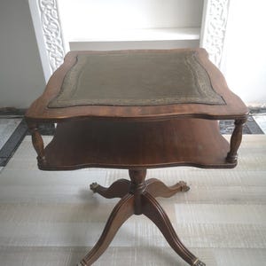 Puede incluir: Una mesa auxiliar de madera vintage con una parte superior cuadrada y un estante inferior. La mesa está cubierta de cuero verde con un borde decorativo. La mesa tiene una base de pedestal con patas de garra. La mesa mide aproximadamente 61 cm de alto.