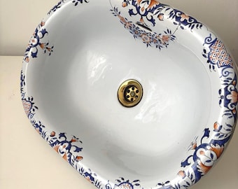 May include: Oval white porcelain sink with a decorative floral border in blue and orange. The sink has a brass drain and a smooth, glossy finish. The floral design is repeated around the rim of the sink.