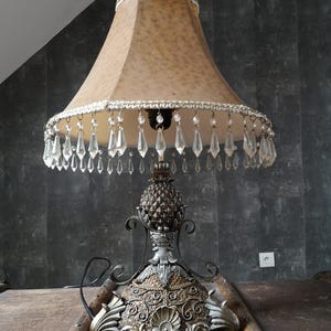 Puede incluir: Una lámpara de mesa con una base con forma de piña y una pantalla beige con flecos de cristal. La lámpara tiene un acabado dorado y plateado.