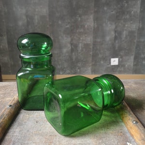 Könnte beinhalten: Zwei grüne Glasbehälter mit Deckeln. Die Behälter sind quadratisch und haben einen abgerundeten oberen Teil. Die Behälter stehen auf einer Holzoberfläche.