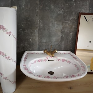 May include: A white porcelain pedestal sink with a floral design and a gold tap. The sink is in a bathroom with a grey tiled wall and a mirror with the word "VOGUE" printed on it.