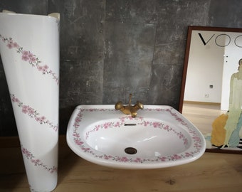 Op de afbeelding: Een wit porseleinen voetwasbak met een bloemenpatroon en een gouden kraan. De wastafel bevindt zich in een badkamer met een grijze betegelde muur en een spiegel met het woord "VOGUE" erop.