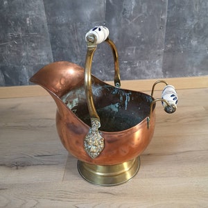 Puede incluir: Un cubo de hielo de cobre y latón con dos asas de porcelana blanca. El cubo tiene un diseño de cabeza de león en la parte delantera y una base redonda.