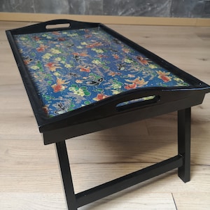 Puede incluir: Una bandeja plegable de madera negra con una parte superior con estampado azul y dorado. La bandeja tiene dos asas y un diseño decorativo de peces y flores.