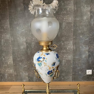 Może przedstawiać: Antyczna lampa mosiężna z białą porcelanową podstawą ozdobioną niebieskimi i złotymi wzorami kwiatowymi. Lampa ma matowy szklany klosz z falbanką na górze.