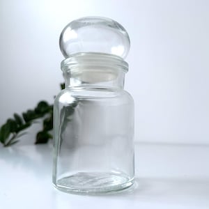 Op de afbeelding: Een heldere glazen apothekerspot met een ronde, heldere glazen stopper. De pot heeft een cilindrisch lichaam en een brede opening. De stopper zit bovenop de pot en zorgt voor een luchtdichte afsluiting. De pot is leeg en staat op een wit oppervlak.