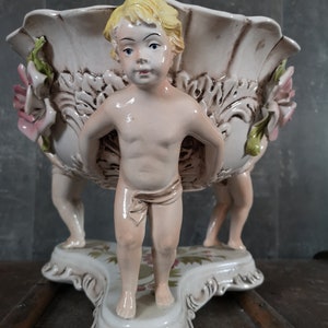 Peut inclure: Un bol en céramique blanche avec un motif floral et trois chérubins qui le tiennent. Les chérubins sont nus, ont les cheveux blonds et des détails peints.
